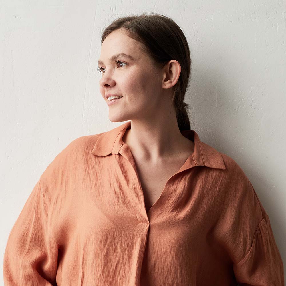 A woman with brown hair wearing a peach shirt looks to the side.
