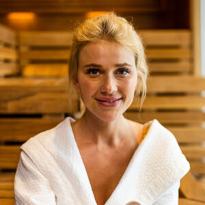 Smiling woman in a white robe inside a wooden sauna.