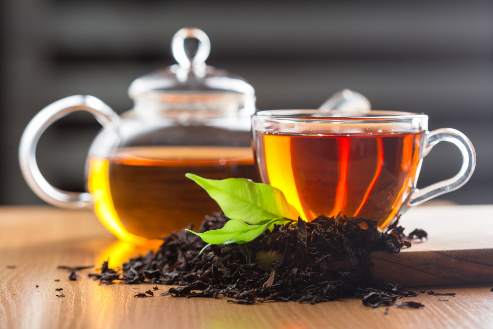 A glass cup and teapot filled with tea beside loose tea leaves and a green leaf.