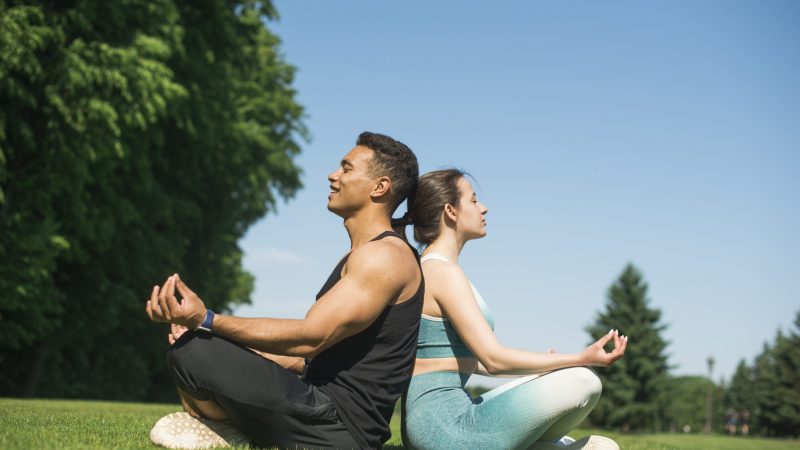 A man and woman meditating back-to-back outdoors on grass.